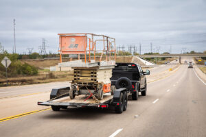 PJ Trailers T6 tilt trailer loaded with a scissor lift, being pulled by a black pickup truck on the freeway